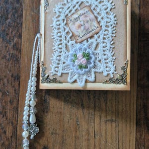 Könnte beinhalten: Handgemachtes Lesezeichen mit quadratischer Karte, weißer Spitze, einem floralen Detail und dem Text "Happiness is Handmade". Es hat eine Perlenquaste mit Kleeblatt-Anhängern. Auf einem Holzgrund.