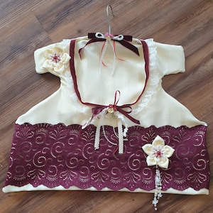 Può includere: Gruccia a forma di vestito color crema con orlo e finiture in pizzo bordeaux. La gruccia presenta un nastro di velluto bordeaux, dettagli in pizzo e abbellimenti floreali decorativi. Un gancio di metallo è in alto.