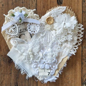 May include: A heart-shaped fabric applique featuring layered white lace, hessian, and crochet elements. It includes a small wooden button, white fabric flowers, a butterfly lace motif with faux pearls, and a measuring tape segment showing 7 inches.