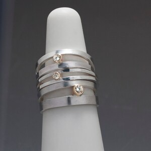 May include: A stack of silver rings with gold accents and small, clear gemstones. The rings are displayed on a white, cylindrical form. The design features a modern, minimalist aesthetic, suitable for everyday wear or special occasions.
