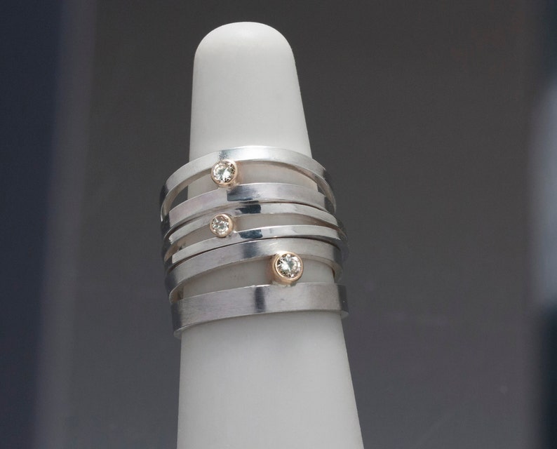 May include: A stack of silver rings with gold accents and small, clear gemstones. The rings are displayed on a white, cylindrical form. The design features a modern, minimalist aesthetic, suitable for everyday wear or special occasions.
