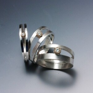 May include: Three silver rings with a unique design. Each ring features a band of silver with a small, round, gold-colored setting holding a clear gemstone. The rings are displayed on a gradient gray surface.