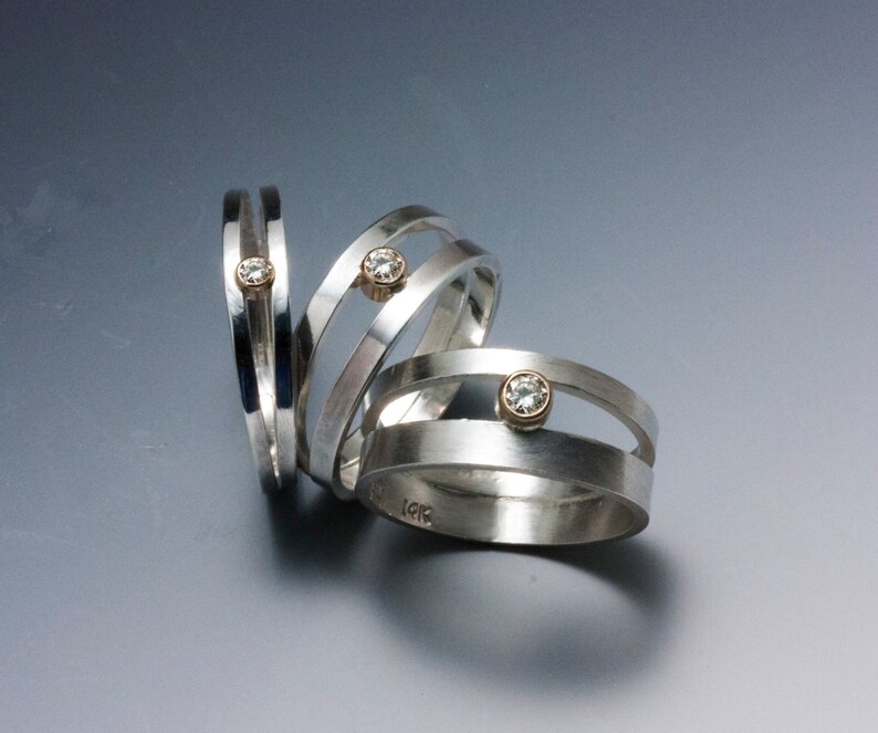 May include: Three silver rings with a unique design. Each ring features a band of silver with a small, round, gold-colored setting holding a clear gemstone. The rings are displayed on a gradient gray surface.