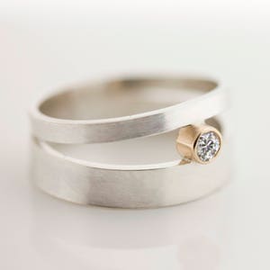 May include: A silver ring with a unique design. The ring features a split band with a gold bezel setting holding a sparkling diamond. The ring is set against a white background, highlighting the contrast between the metals and the diamond.