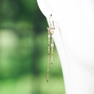 May include: A gold-colored earring with a delicate chain design. The earring features a small bar at the top, with a series of elongated oval and small cylindrical links that dangle down. The background is a soft green and white.