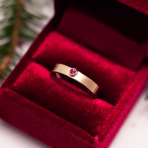 May include: A gold ring with a single red gemstone set in the band, displayed in a red velvet ring box.