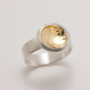 Puede incluir: Un anillo de plata con una banda ancha y un centro circular dorado. El centro dorado tiene una apariencia texturizada y martillada, engastado en un bisel plateado. El anillo se muestra sobre un fondo blanco.