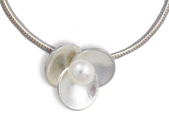May include: A silver necklace featuring a floral pendant with three overlapping, textured, circular petals. A single, round, white pearl sits in the center of the petals. The necklace has a silver, snake chain.
