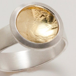 Puede incluir: Un anillo de plata con una piedra central dorada y texturizada. El anillo tiene una banda ancha y un engaste circular alrededor del elemento dorado. El centro dorado tiene una superficie rugosa y desigual.