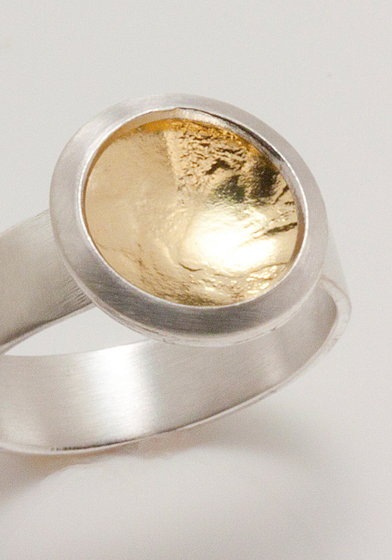 Puede incluir: Un anillo de plata con una piedra central dorada y texturizada. El anillo tiene una banda ancha y un engaste circular alrededor del elemento dorado. El centro dorado tiene una superficie rugosa y desigual.