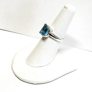 May include: A silver ring featuring a large, rectangular, light blue gemstone. The gemstone is set in a simple, polished silver band. The ring is displayed on a white ring holder, showcasing its design and color.