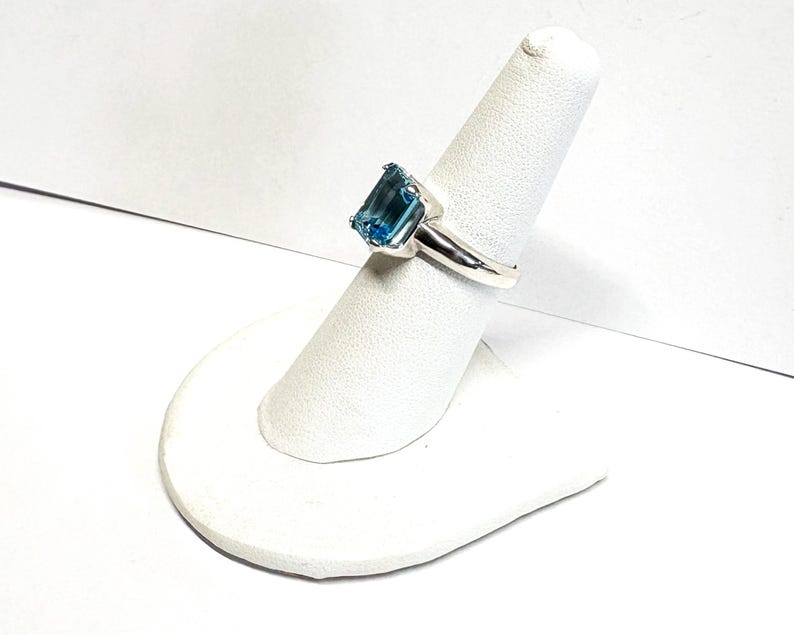May include: A silver ring featuring a large, rectangular, light blue gemstone. The gemstone is set in a simple, polished silver band. The ring is displayed on a white ring holder, showcasing its design and color.