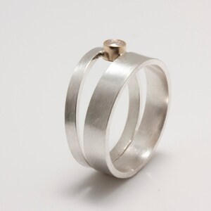 May include: A modern silver ring with a unique design. The ring features three bands, with one band extending upwards to hold a small, gold-colored accent. The ring is set against a white background.
