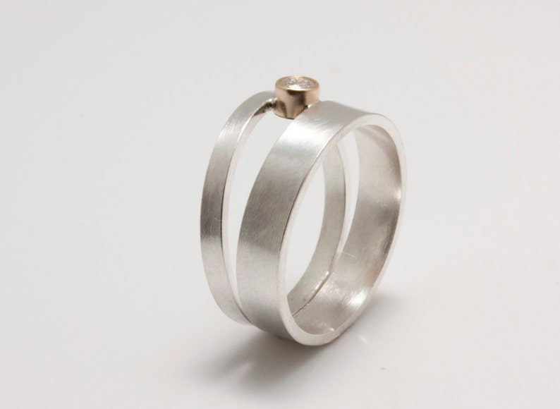 May include: A modern silver ring with a unique design. The ring features three bands, with one band extending upwards to hold a small, gold-colored accent. The ring is set against a white background.