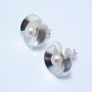 May include: A pair of silver earrings with a pearl in the center of each earring. The earrings are shaped like a flower with a curved petal design.