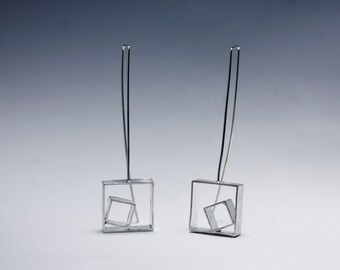 May include: A pair of silver earrings with a geometric design. Each earring features a square frame with a smaller square inside, creating a layered effect.