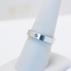 May include: A silver ring with a single, small, light blue gemstone. The ring is set on a white display stand, showcasing its simple, elegant design. The band is flat and wide, with a brushed metal finish.