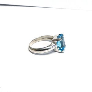 May include: A silver ring featuring a large, rectangular, light blue gemstone. The ring has a simple, elegant design with a polished finish. The gemstone is the focal point, catching the light and adding a pop of color.