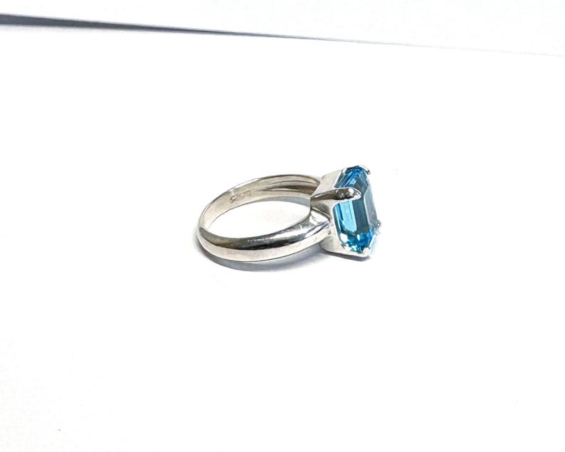 May include: A silver ring featuring a large, rectangular, light blue gemstone. The ring has a simple, elegant design with a polished finish. The gemstone is the focal point, catching the light and adding a pop of color.