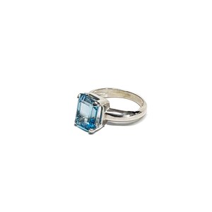 May include: A silver ring featuring a large, rectangular, light blue gemstone. The gemstone is the focal point, set in a simple, polished silver band. The ring is displayed against a white background, highlighting its clean lines and color.