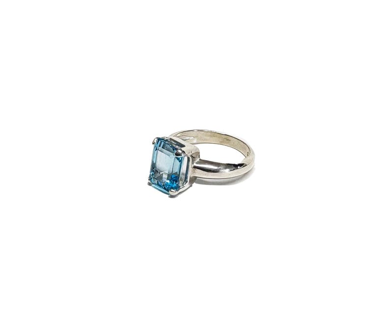 May include: A silver ring featuring a large, rectangular, light blue gemstone. The gemstone is the focal point, set in a simple, polished silver band. The ring is displayed against a white background, highlighting its clean lines and color.