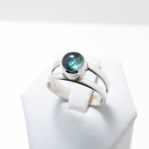 May include: A silver ring featuring a round, cabochon-cut teal blue gemstone set in a smooth bezel. The band is constructed with two parallel silver wires that meet at the base, creating an open, split-shank design with a polished metallic finish.