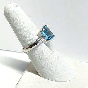 May include: A silver ring featuring a large, rectangular, light blue gemstone. The gemstone is set in a simple, polished silver band. The ring is displayed on a white, textured finger form, showcasing its design and color.