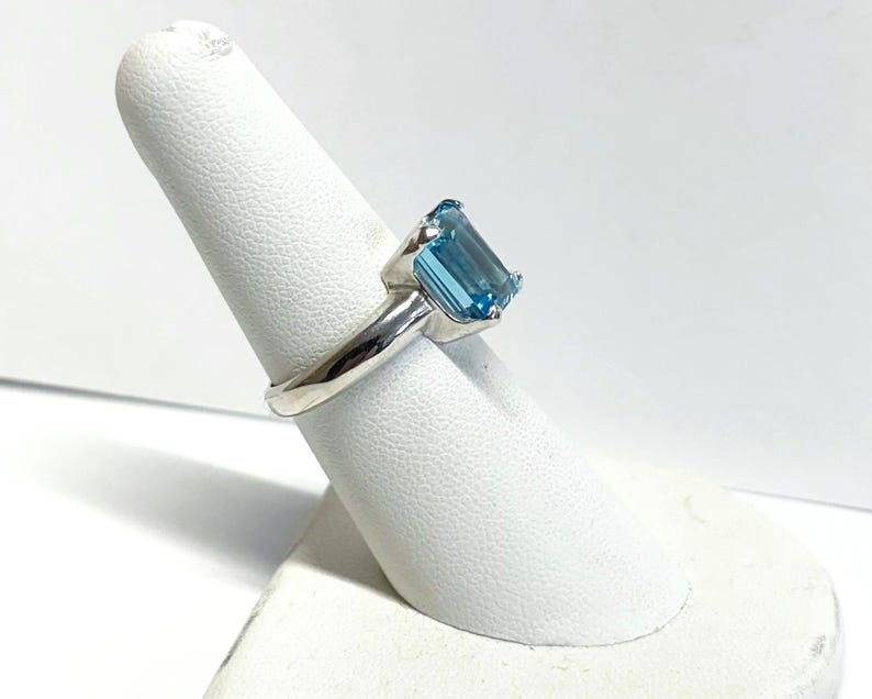 May include: A silver ring featuring a large, rectangular, light blue gemstone. The gemstone is set in a simple, polished silver band. The ring is displayed on a white, textured finger form, showcasing its design and color.