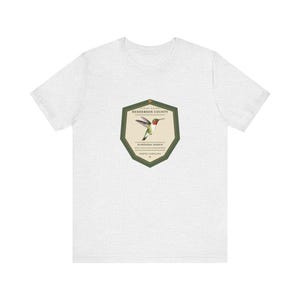 May include: A light heather gray short-sleeve t-shirt featuring a central shield-shaped graphic. The emblem displays a hummingbird illustration with the text FIELD GUIDE, HENDERSON COUNTY, Archilochus colubris, RUBY-THROATED HUMMINGBIRD, NORTH CAROLINA.