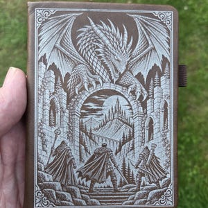 May include: A brown leather-bound journal featuring a detailed laser-engraved illustration of a large dragon perched above a stone archway. Below the arch, three figures stand facing a distant castle, surrounded by pine trees and rocky terrain.