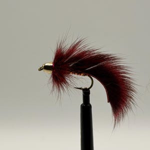 May include: A fishing fly lure featuring deep burgundy fur fibers extending from a metallic gold bead head. The lure is constructed on a curved metal hook with a sharp point, showcasing a textured body and long, flowing dark red hair-like material.