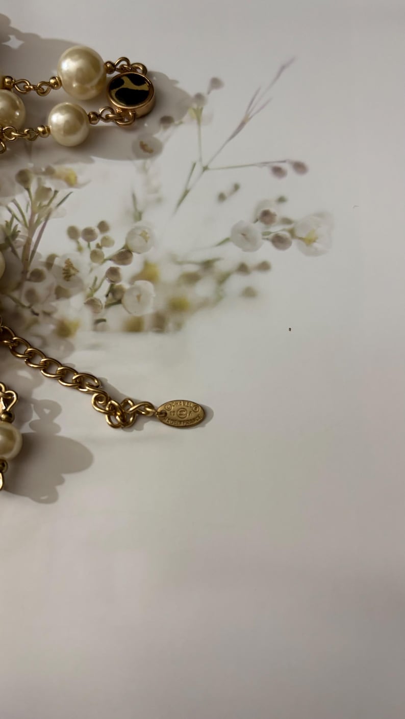 Puede incluir: Un collar de cadena en tono dorado con perlas de imitaci&oacute;n color crema y detalles circulares en negro y dorado. El collar incluye una peque&ntilde;a etiqueta met&aacute;lica ovalada grabada con el logo de Chanel y el texto Made in France en los eslabones.