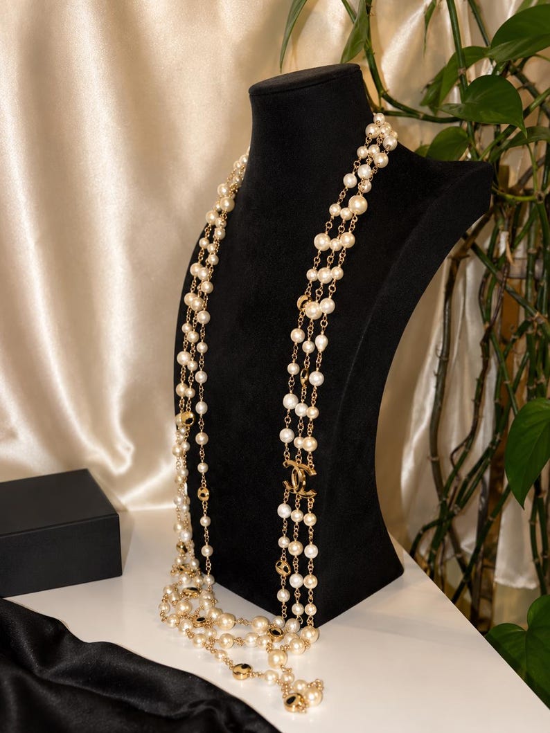 Puede incluir: Un collar largo de varias hebras con perlas artificiales de color crema intercaladas con eslabones de metal dorado y detalles circulares. El dise&ntilde;o incluye un emblema central de metal dorado con el logotipo CC entrelazado.