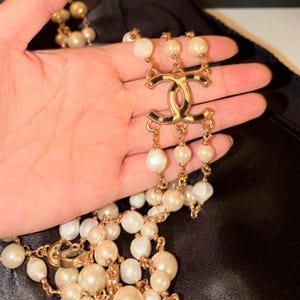 Puede incluir: Un collar de m&uacute;ltiples hilos con perlas blancas brillantes intercaladas con cadenas de metal dorado. La pieza central es un prominente emblema dorado con el logotipo CC entrelazado y detalles en negro, que conecta tres hilos de perlas.