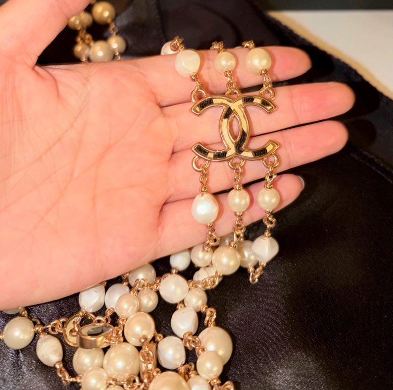 Puede incluir: Un collar de m&uacute;ltiples hilos con perlas blancas brillantes intercaladas con cadenas de metal dorado. La pieza central es un prominente emblema dorado con el logotipo CC entrelazado y detalles en negro, que conecta tres hilos de perlas.