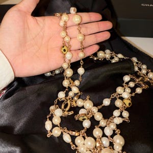 Puede incluir: Un collar de cadena larga en tono dorado con m&uacute;ltiples perlas blancas de varios tama&ntilde;os y detalles circulares dorados. El collar incluye un destacado dije dorado con el logotipo CC entrelazado integrado en el dise&ntilde;o.