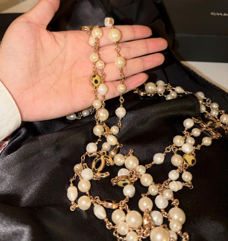 Puede incluir: Un collar de cadena larga en tono dorado con m&uacute;ltiples perlas blancas de varios tama&ntilde;os y detalles circulares dorados. El collar incluye un destacado dije dorado con el logotipo CC entrelazado integrado en el dise&ntilde;o.