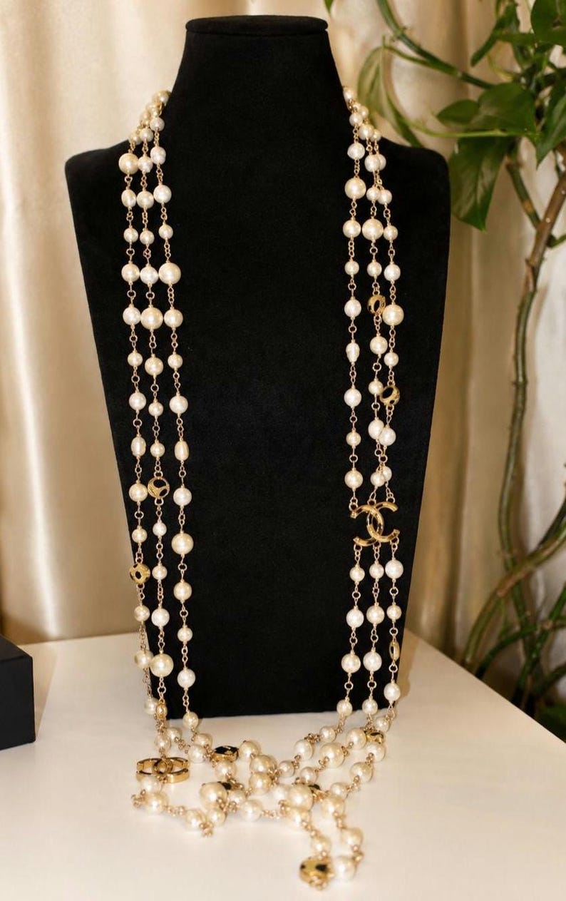 Puede incluir: Un collar largo de varias hebras con perlas blancas redondas intercaladas con eslabones de cadena de metal dorado. El dise&ntilde;o incluye dos logotipos CC entrelazados de metal dorado integrados en las hebras, creando un aspecto en capas.