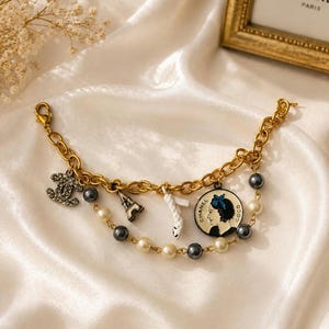 May include: A gold-tone chain bracelet featuring multiple charms including a crystal-encrusted interlocking logo, a miniature Eiffel Tower, a white shoe, and a circular pendant depicting a profile silhouette with the text CHANEL COCO.