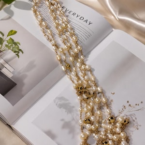 Puede incluir: Un collar largo de varias hebras con perlas de color crema y cuentas de metal dorado pulido. El collar est&aacute; acentuado con un dije central de logo CC entrelazado en tono dorado, con las hebras conectadas por delicadas cadenas de metal dorado.