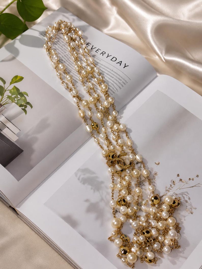 Puede incluir: Un collar largo de varias hebras con perlas de color crema y cuentas de metal dorado pulido. El collar est&aacute; acentuado con un dije central de logo CC entrelazado en tono dorado, con las hebras conectadas por delicadas cadenas de metal dorado.