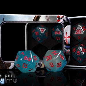 May include: A set of translucent blue twenty-sided dice with red numbering, housed in a rectangular metal tin featuring graphic artwork and the text "designed for CORVUS BELLI INFINITY". Two dice are placed in front of the open tin.