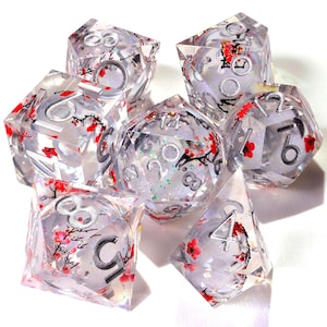 May include: A set of seven polyhedral dice made of clear resin, each featuring embedded red cherry blossom branch illustrations and silver-colored numbers. The dice have sharp edges and contain subtle iridescent glitter particles throughout.