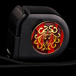 Peut inclure: Un mètre ruban rétractable noir doté d'un emblème circulaire doré et rouge sur le côté. L'emblème représente un visage stylisé aux cheveux en forme de serpents dorés sur un fond rouge foncé. Une dragonne noire est fixée sur le côté.