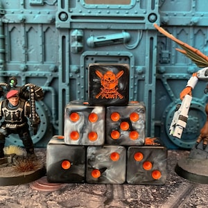 May include: A stack of six-sided dice with a black and white marbled finish and bright orange pips. The top die features an orange graphic of a helmeted skull and the word POINT. The dice are positioned between two painted miniature game figures.