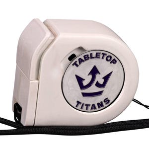 May include: A white retractable measuring tape with a black wrist strap. The side features a circular emblem with a purple crown graphic and the text "TABLETOP TITANS" printed in purple. The device has a smooth, rounded plastic casing.