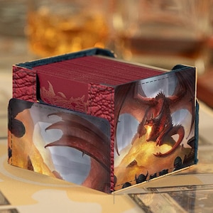 May include: A rectangular deck box for trading cards featuring a textured red exterior and a printed graphic of a red dragon perched on a pile of gold coins. The box is open at the top, revealing a stack of red-backed playing cards inside.