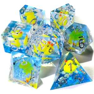 May include: A set of seven polyhedral dice made of clear blue resin, each containing a miniature yellow rubber duck and small white cloud-like inclusions. The dice feature silver-colored numbers engraved on the faces, including a d20, d12, d10, d8, d6, and d4.