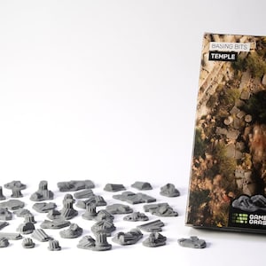 May include: A collection of gray miniature temple pieces and a box with a temple scene. The box has the text "BASING BITS TEMPLE" and "GAMERS GRASS". The miniature pieces are scattered on a white surface.
