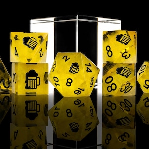 May include: A set of seven translucent yellow polyhedral dice with black numbering and a black beer mug icon on the highest face of each die. The dice are arranged in a group against a black background with a clear acrylic block behind them.
